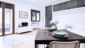 A modern dining area with a dark wooden table set for two, white chairs, and a green bowl centerpiece. A white cabinet with a flat-screen TV is visible in the background. - Easylife - Studio with balcony in Risorgimento (Milano)