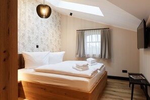 Une chambre avec un lit en bois, une lucarne, un luminaire au plafond, une fenêtre avec des rideaux et une petite table sur laquelle se trouve un magazine.
