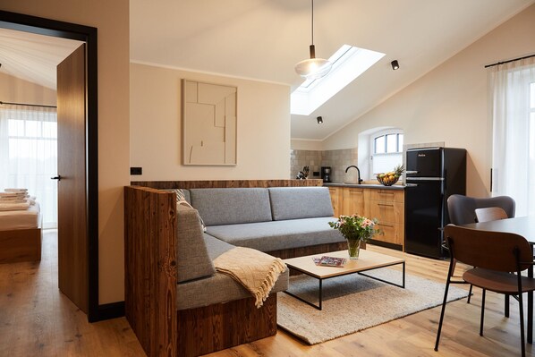 Un salon moderne avec un canapé gris, des éléments décoratifs en bois, une table basse avec un magazine, et un coin cuisine comprenant un réfrigérateur et un évier.