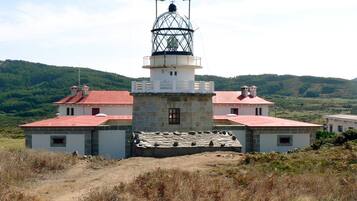 Un faro con base de piedra y techo rojo, rodeado por un sendero de tierra y colinas verdes al fondo.