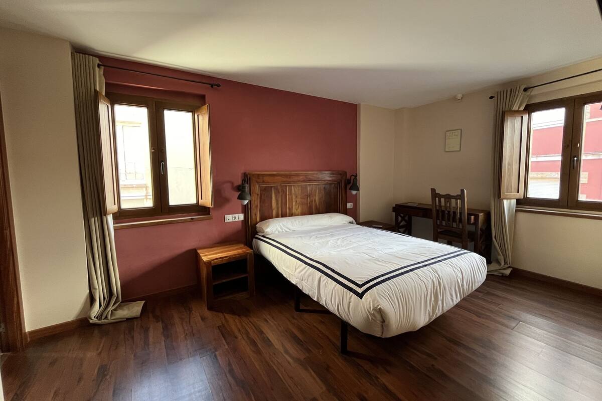Un dormitorio con una cama de madera, una mesita de noche, un escritorio y dos ventanas.
