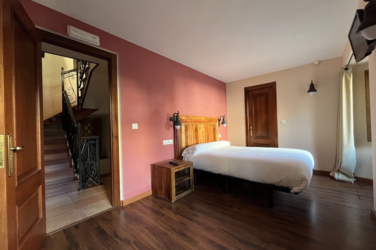 Un dormitorio con suelo de madera, una cama, una mesita de noche, cabecera, una puerta y una escalera.