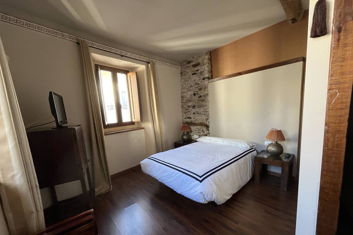 Un dormitorio con cama, suelo de madera, pared de piedra, ventana y lámparas de noche.