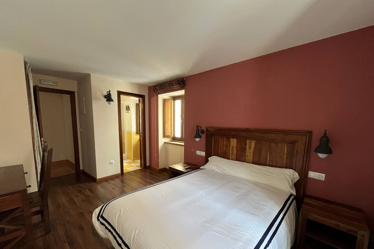 Un dormitorio con una cama grande, suelo de madera, una pared de acento roja y una ventana con vistas al pasillo.
