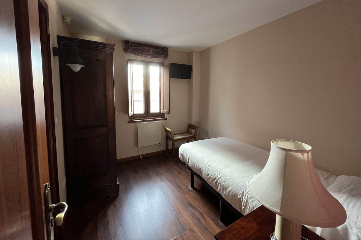 Un dormitorio pequeño y ordenado con una cama individual, una silla, un escritorio y una ventana con cortinas.