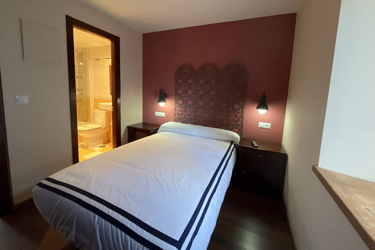 Una cama bien hecha en una habitación de hotel, con cabecera, dos lámparas de noche y una mesita de noche de madera.