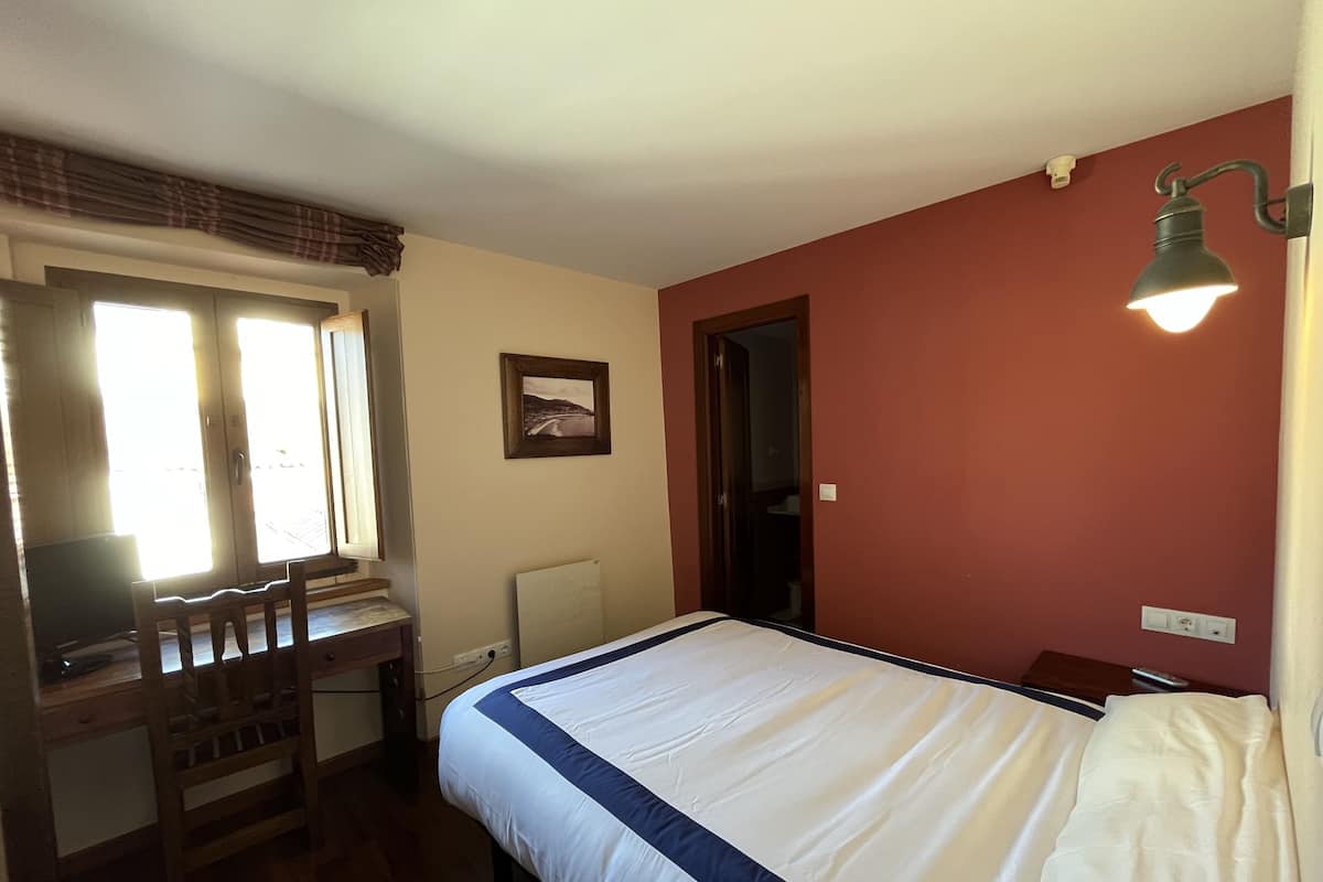Un dormitorio con cama, escritorio, silla, ventana con cortinas, puerta y lámpara fijada a la pared.