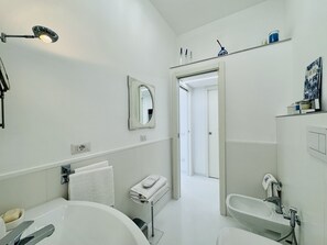 Ein modernes Badezimmer mit einer Badewanne, einem Waschbecken und einem Spiegel. Handtuchhalter und ein Ablagefach mit Pflegeprodukten sind ebenfalls vorhanden.