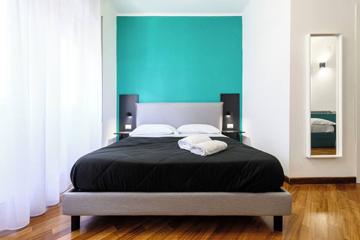 A modern bedroom with a teal accent wall, a large bed with a grey headboard, two bedside tables with lamps, and a mirror on the wall.
