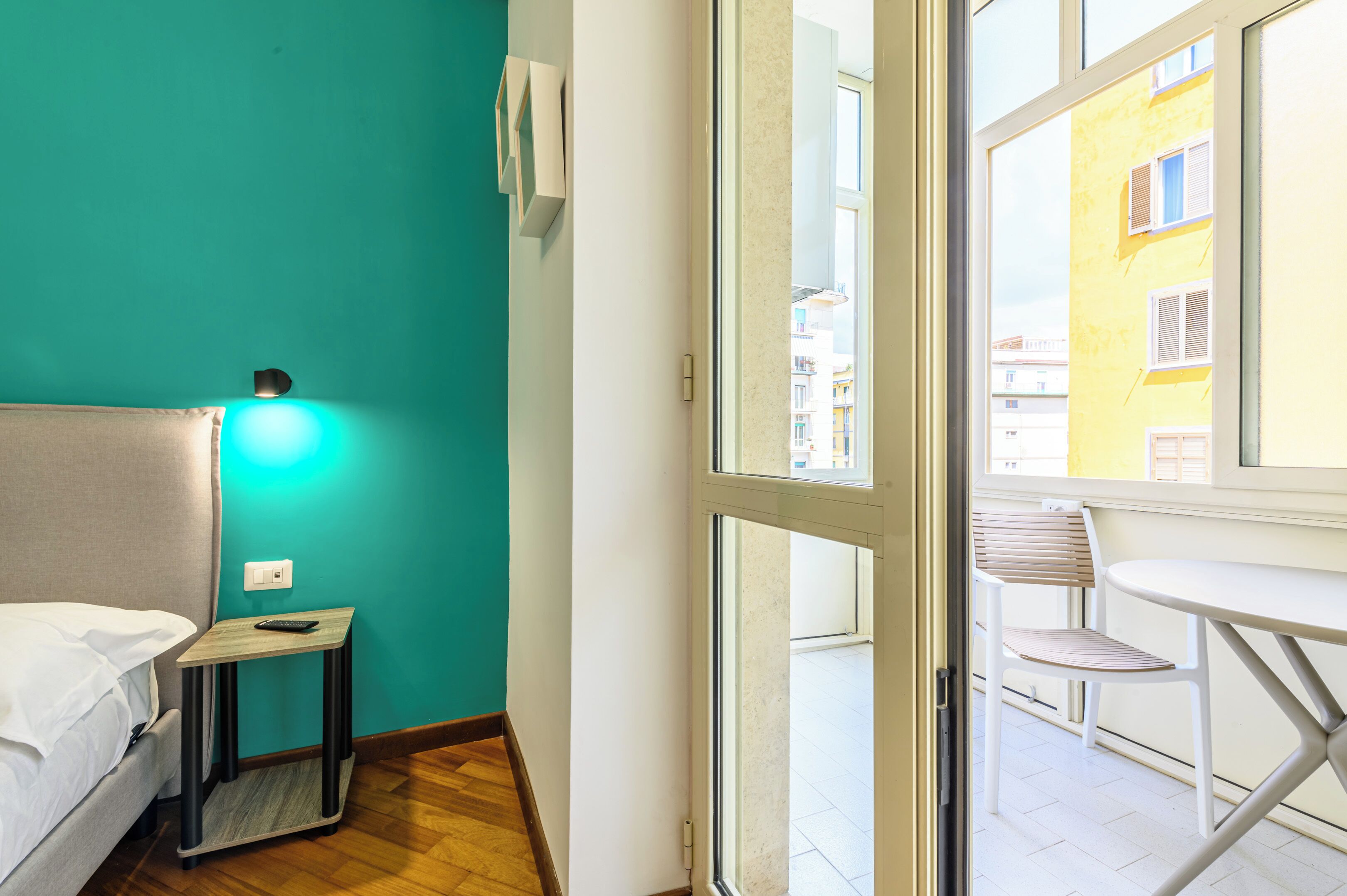 Habitación de hotel con cama, una mesita con control remoto, toma de corriente y ventana que da a un balcón exterior con una silla y una mesa.