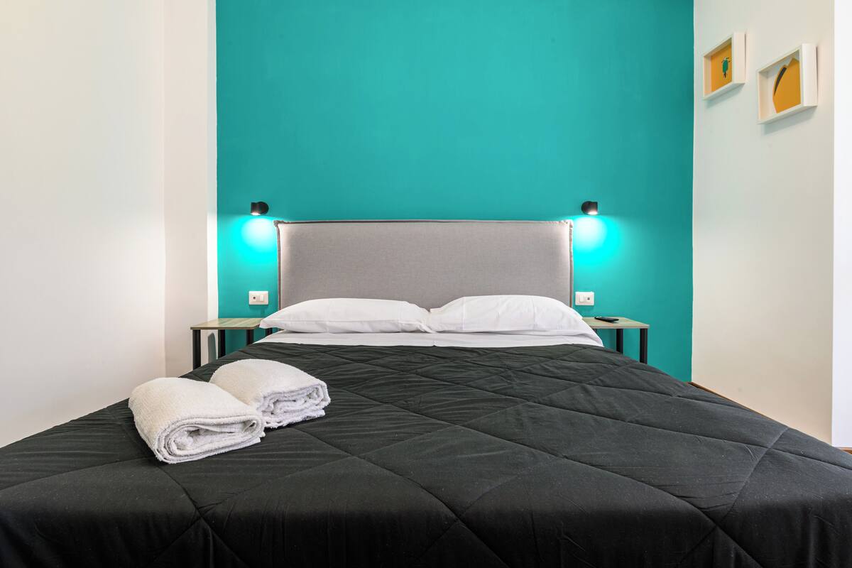 A neatly made bed with a black quilt, two white pillows, and two folded towels on the side.