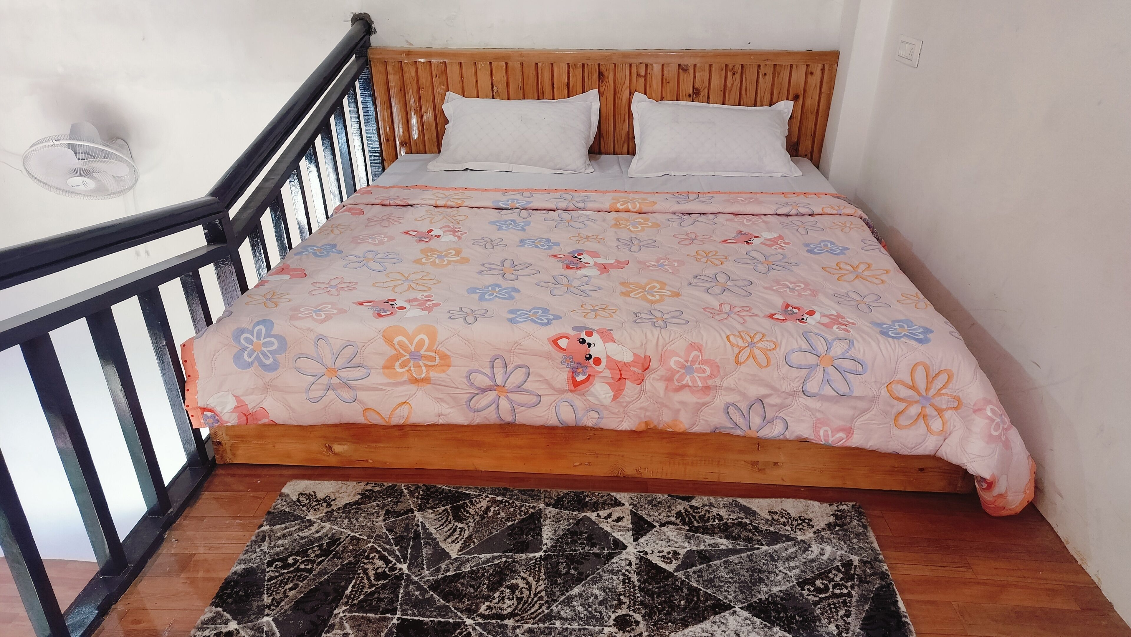 A bedroom with a wooden bed, two pillows, a floral bedspread, a black and white rug, and a ceiling fan.