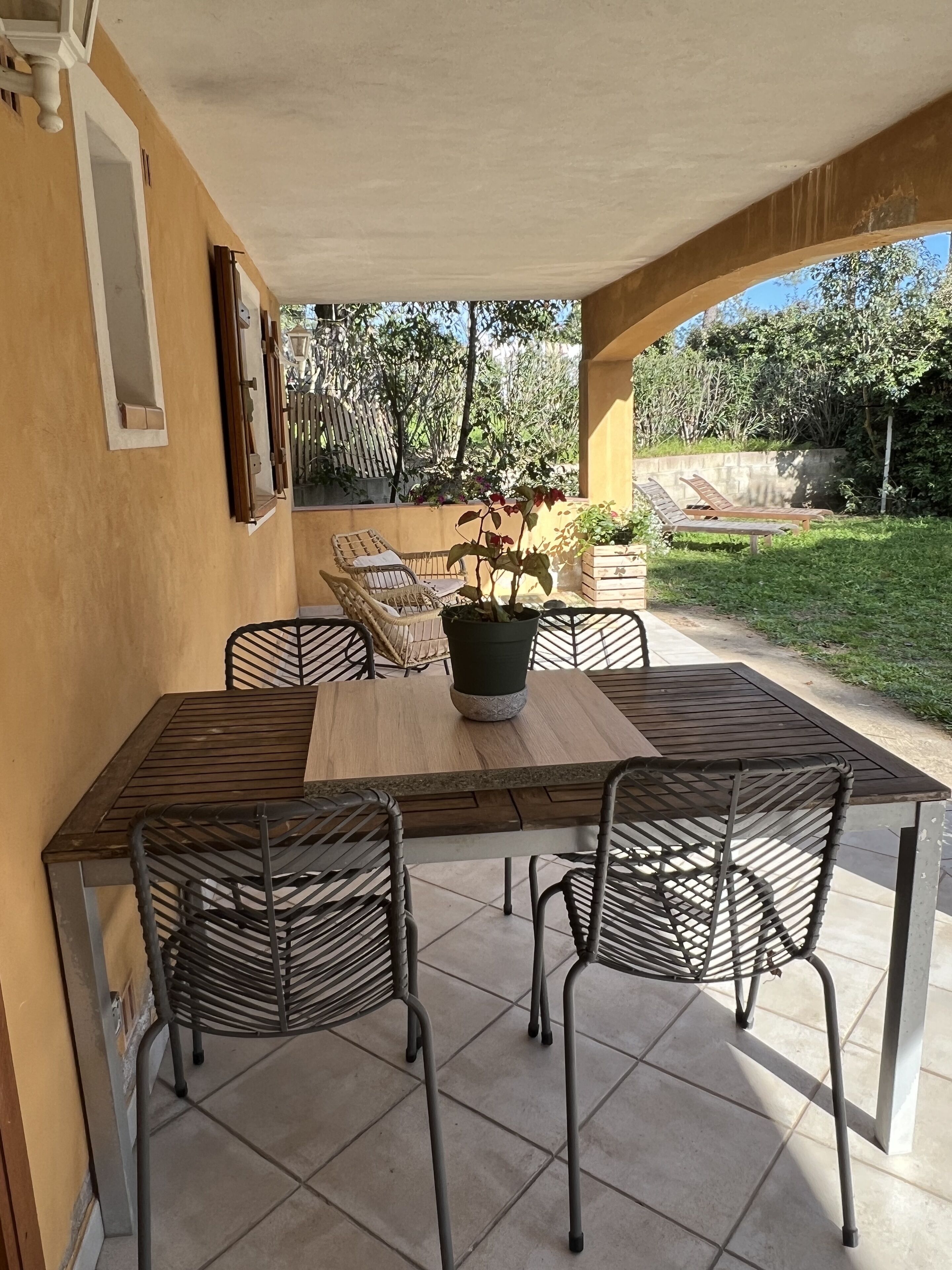 A covered patio area with a wooden table and four chairs, surrounded by greenery and a view of trees.