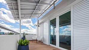 A covered outdoor area with a wooden floor, white walls, and a glass door leading to a balcony. There are two white chairs and a large blue planter with green plants. - Beach Condo by Otium | Steps from Oceanpark Beach (San Juan)