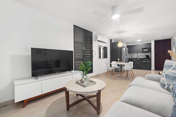 A modern living room with a flat-screen TV, a round coffee table, a sofa, and a dining area with chairs.