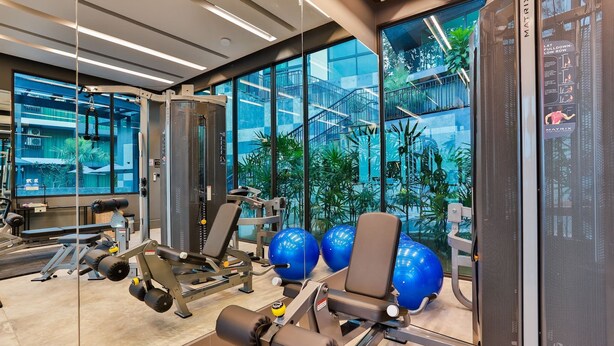 Une salle de sport moderne équipée de matériel d’exercice, dotée de grandes fenêtres et offrant une vue sur un bâtiment avec des escaliers et de la verdure.
