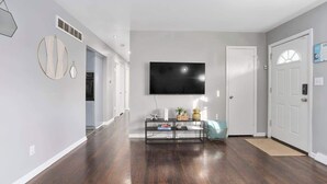 Un vaste salon doté d’un téléviseur à écran plat fixé au mur, d’un parquet en bois foncé et d’une porte blanche située à droite.