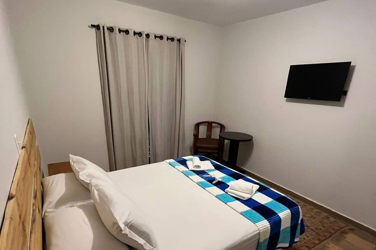 A single bed with a wooden headboard, a bedside table, a chair, a flat-screen TV mounted on the wall, and a window with curtains.