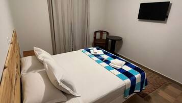 A single bed with a wooden headboard, a bedside table, a chair, a flat-screen TV mounted on the wall, and a window with curtains.
