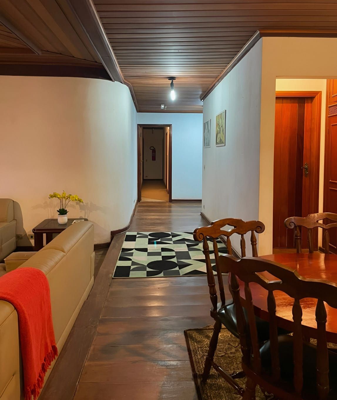 A hallway leading to a dining area with a patterned rug, a sofa with a red throw, and a wooden dining table with chairs.