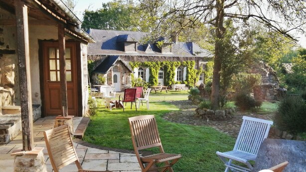 A patio with wooden chairs and a stone table, a stone wall, and a building with a thatched roof in the background. - Charming Country Retreat: Sleeps 11, Indoor Pool, Pet-Friendly, Near Château-Gontier (Gennes-Longuefuye)