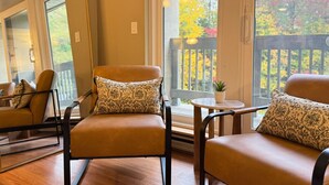 A cozy seating area with leather chairs and patterned cushions, a small round table with a potted plant, and a view of a wooden deck through large windows. - CountryCub- Pool, Hot Tub, Gym, Sauna, Near Resort (Killington)