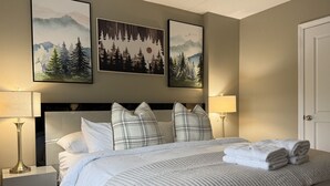 A neatly made bed with white bedding and plaid pillows, flanked by bedside tables with lamps, in a room with a neutral color scheme and framed artwork on the walls. - CountryCub- Pool, Hot Tub, Gym, Sauna, Near Resort (Killington)