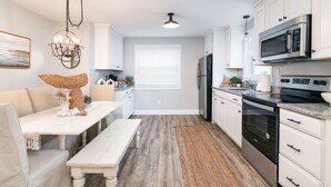 A modern kitchen with white cabinetry, stainless steel appliances, and a dining area with a white table and bench. - Hidden Gem in N. OC on the Beach (Ocean City)