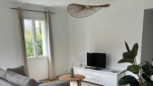 Een moderne woonkamer met een grijze bank, een ronde houten salontafel, een flatscreen televisie op een wit kastje, en een grote plant in een pot.
