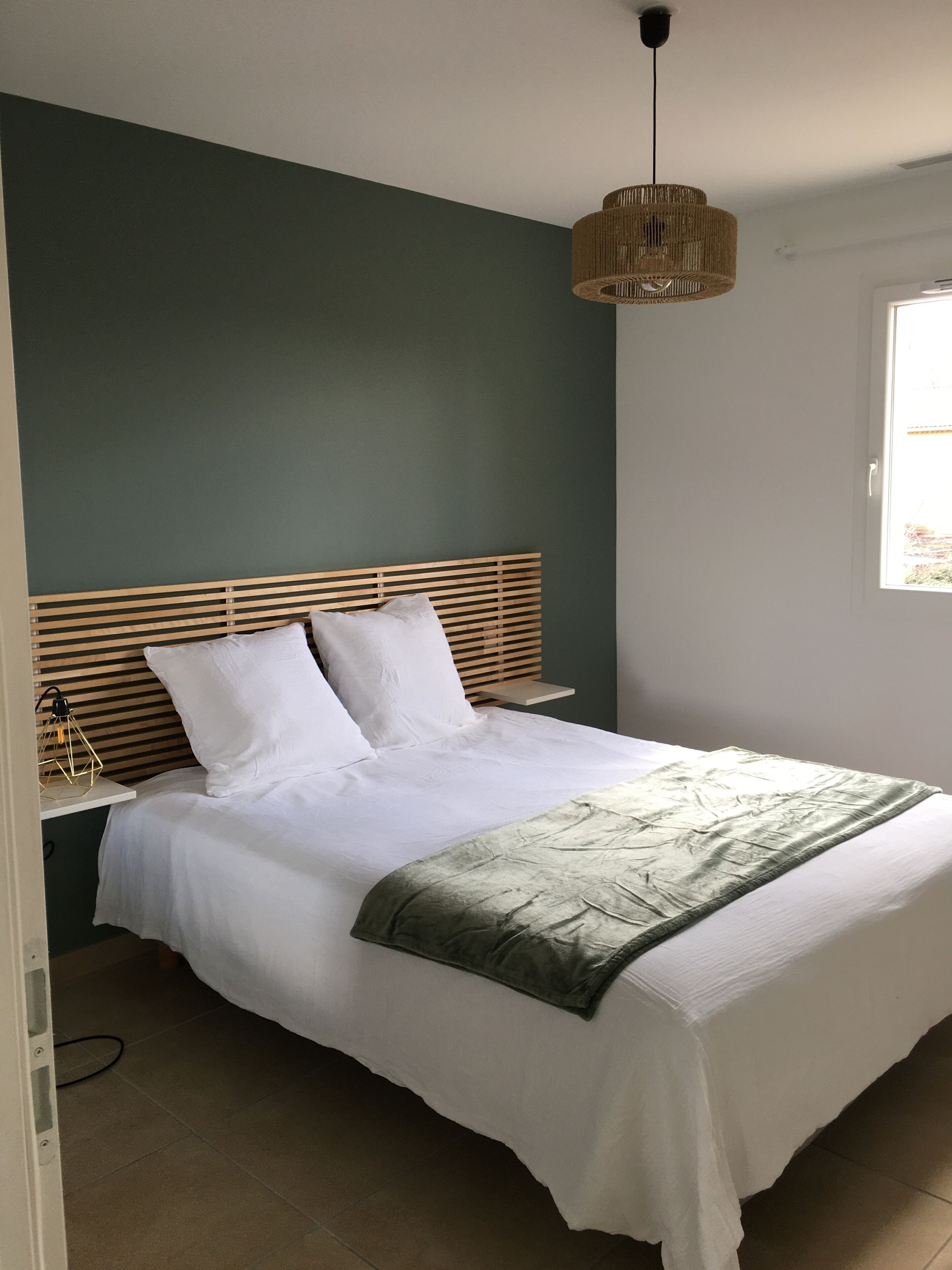 Un lit bien fait, avec du linge de lit blanc et un plaid vert, une tête de lit en bois et un luminaire suspendu.