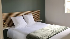 Een net opgemaakt bed met witte lakens, een groene plaid, een houten hoofdbord en een hangende lamp.