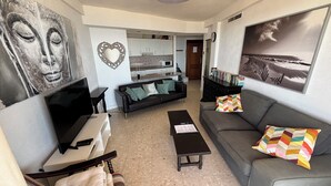 Una sala de estar con televisor, sofá, mesa de centro y una cocina al fondo.