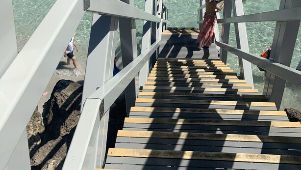 Gescales de fusta que condueixen a un moll sobre aigües turqueses cristal·lines, amb una persona amb camisa blanca i pantalons curts negres que hi baixa.