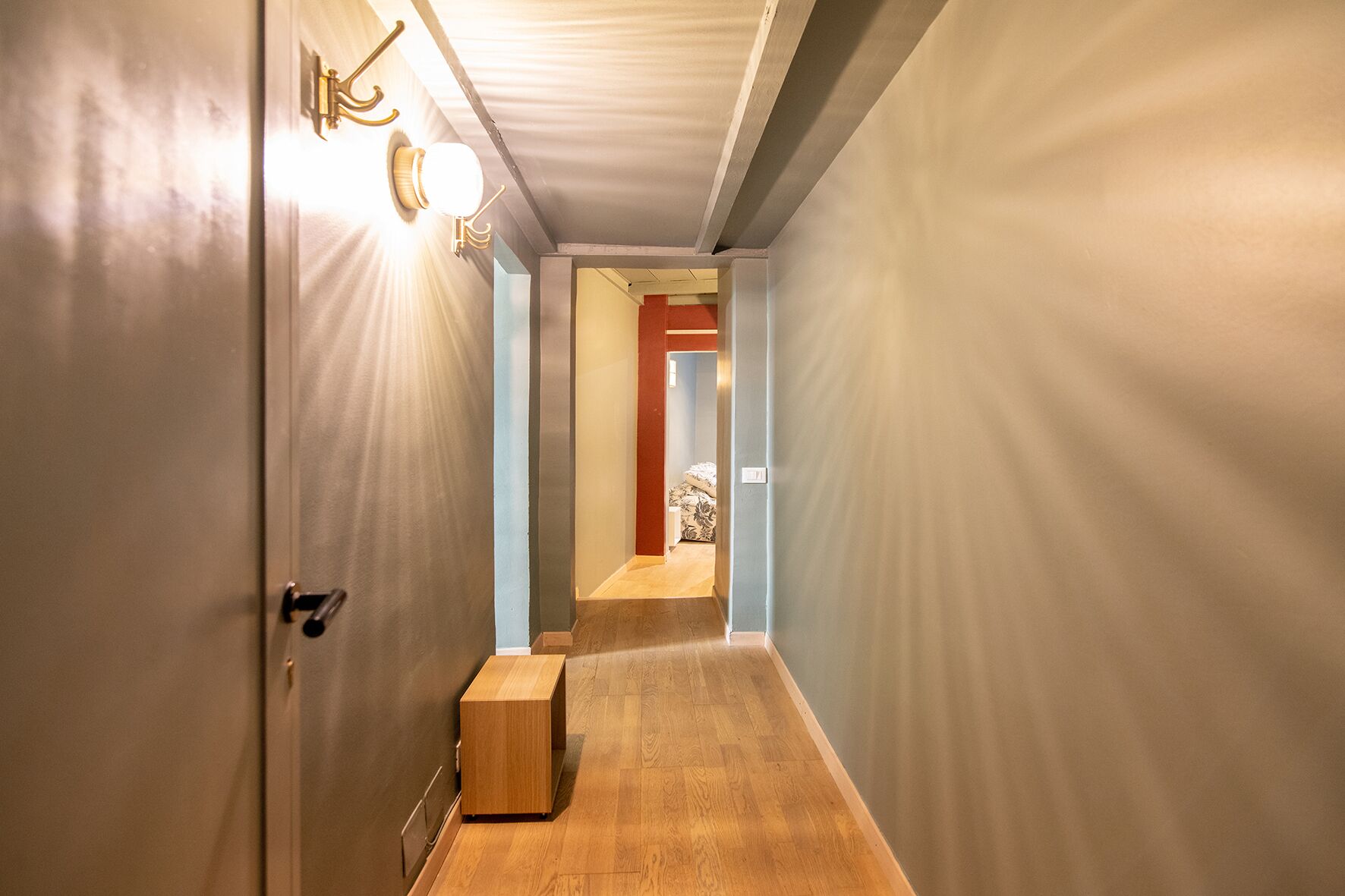 A narrow hallway with a wooden bench, a door with a light fixture, and a visible room in the distance.
