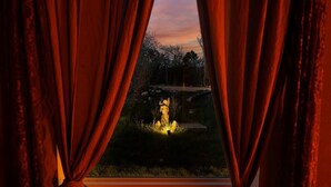 Una ventana con cortinas, vista de una fuente iluminada y un sendero que lleva a un edificio.