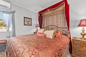 A four-poster bed with a red canopy, a wooden nightstand with a lamp, and a window with curtains. - The Kuhl Little Castle Haus (Fredericksburg)