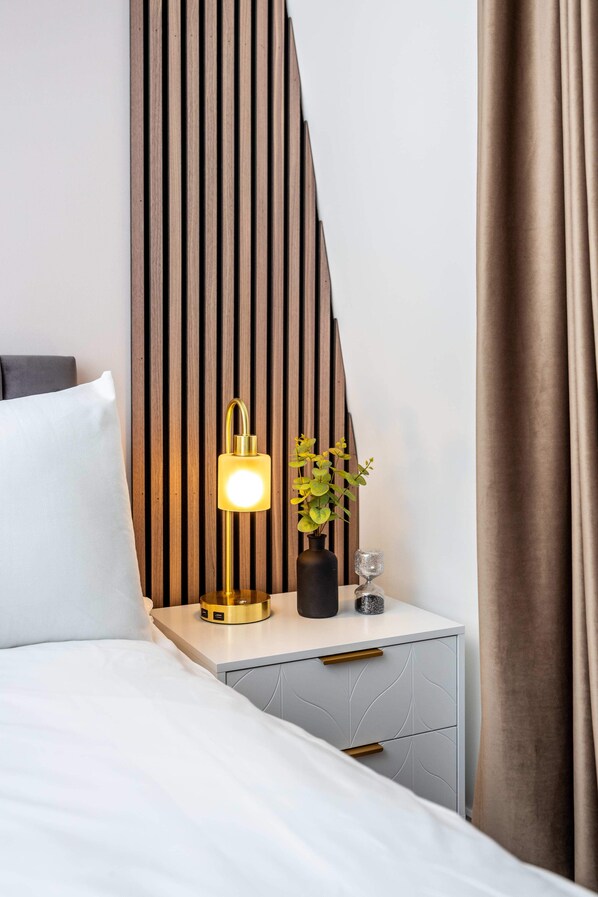 A neatly made bed with white linens, a bedside lamp, a small potted plant, and a decorative glass on a bedside table.