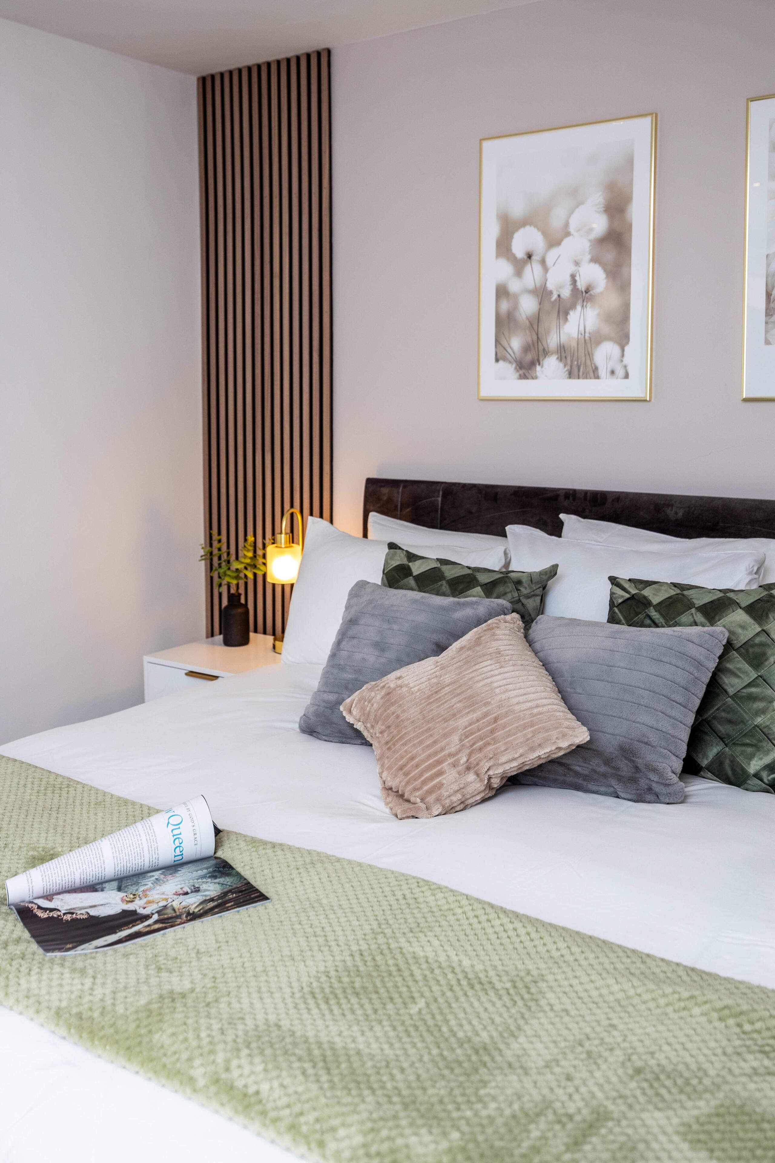 Un lit bien fait, recouvert d’une couverture verte, une table de chevet avec une lampe, et deux tableaux encadrés accrochés au mur.