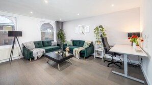 Un salon moderne avec un canapé vert, une table basse, un bureau avec une chaise et une grande fenêtre donnant sur des immeubles.