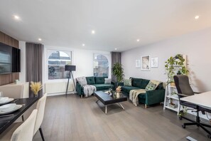 Un salon moderne avec un canapé, une table basse et un bureau avec une chaise. On y trouve des fenêtres, une lampe et une étagère pour les plantes.