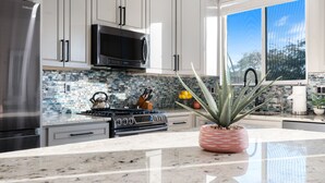 A modern kitchen with white cabinets, a stainless steel refrigerator, a black microwave, a gas stove, and a large window offering a view of trees. - 3BD - Heated Pool - Pet friendly - N. Scottsdale (Cave Creek)