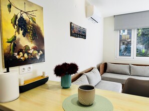 Un salon avec un tableau représentant des grappes de raisin, un vase avec des fleurs rouges, une tasse à café et une fenêtre donnant sur des immeubles.
