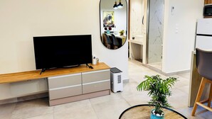 Een moderne woonkamer met een flatscreen televisie, een houten dressoir, een spiegel, een eethoek met stoelen, een koelkast en een potplant.