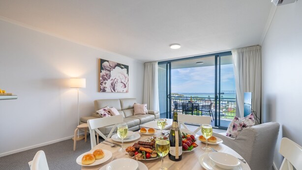 A modern hotel room with a balcony, a dining table set with food and drinks, a sofa, and a painting on the wall. - The Landmark unit 405 Resort style, spectacular water views, lift, linen, pool, games room, port-a-cot and highchair. (Nelson Bay)