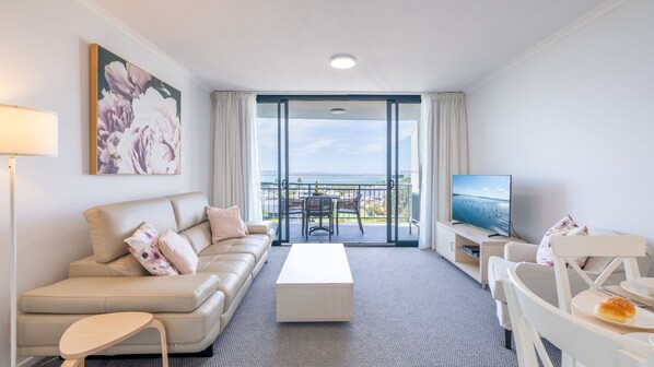 A modern hotel room with a balcony, a sofa, a coffee table, a TV, and a dining area.