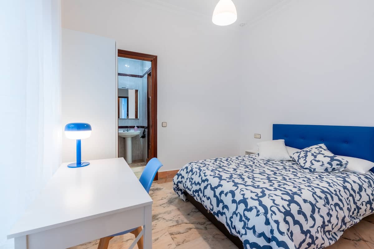 A bedroom with a bed, a desk, a chair, and a bathroom visible through an open door.