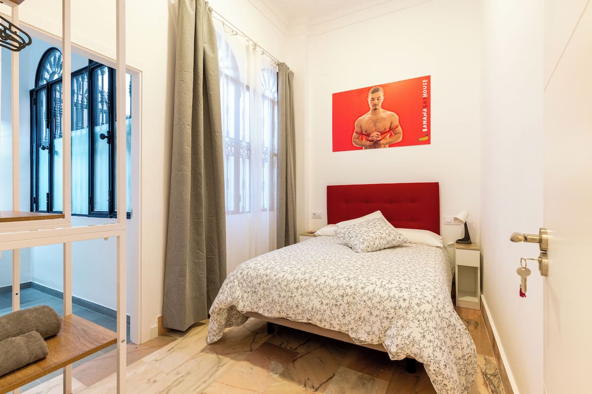 A bedroom with a bed, a nightstand, a red headboard, a window with curtains, and a poster on the wall.