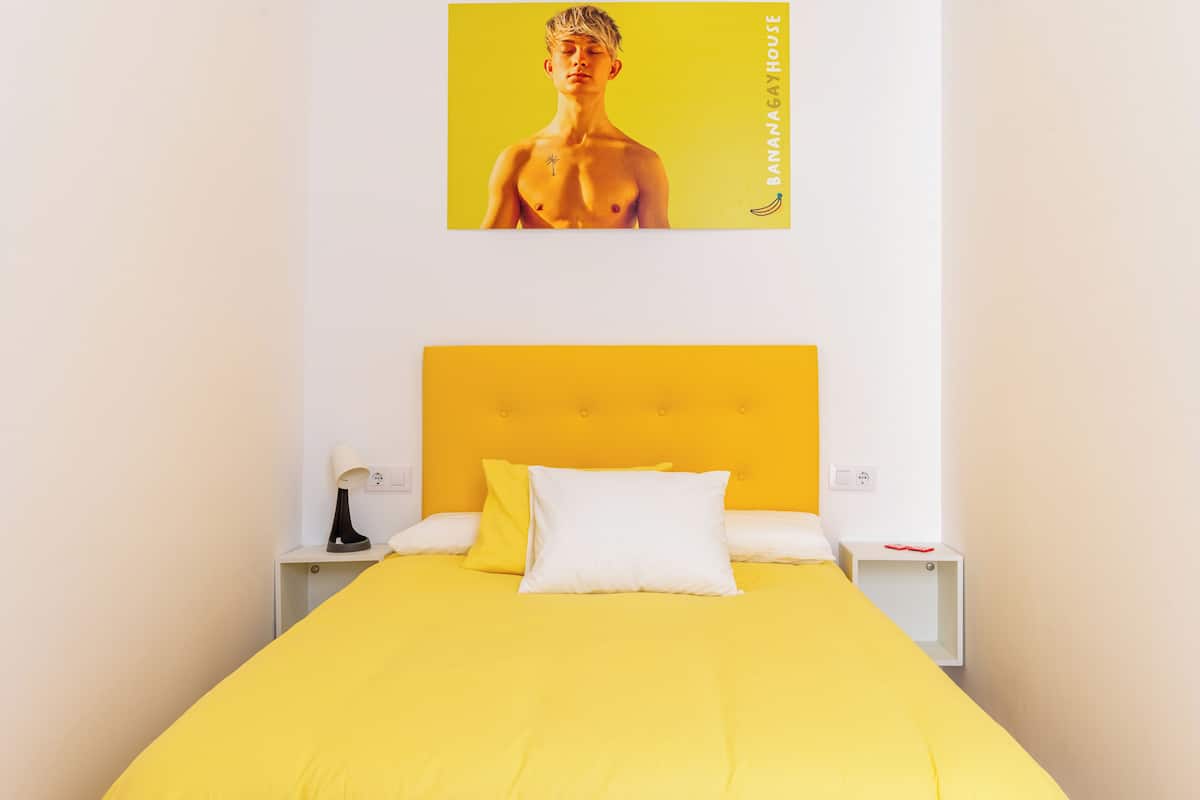 A bed with a yellow headboard and white bedding, a nightstand with a lamp, and a wall-mounted picture.