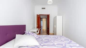 Un dormitorio con una cabecera morada, una colcha estampada, un almohadón blanco y un escritorio blanco con una silla.