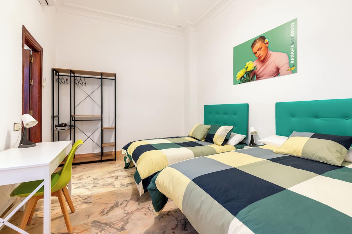 A bedroom with two beds, a desk, a chair, a wardrobe, and a wall-mounted picture.